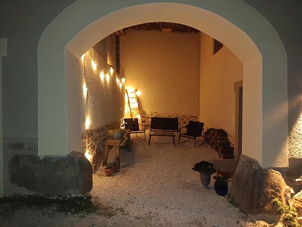 An outdoor seating area with stone walls, a wooden table, and chairs, illuminated by string lights.