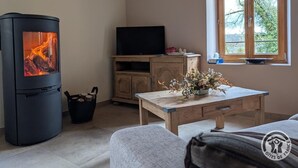 A cozy living room with a wood-burning stove, a television on a wooden cabinet, a window with a view of trees, and a table with a vase of flowers. - "Ensemble - MEYS - Gîte (MEYS)