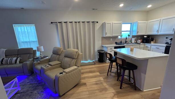 A modern living room with a beige sofa, a small table, and a coffee table. There is a kitchen area with white cabinets, a sink, and a stove. A window with curtains is visible in the background.