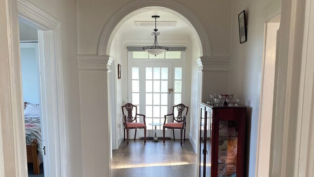 A hallway with wooden flooring, white walls, and an arched doorway leading to a room with a bed. There are two chairs and a small table in the room.