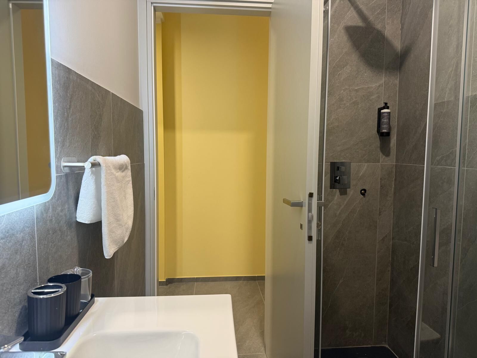 Ein Badezimmer mit weißem Waschbecken, einem Handtuchhalter mit Handtuch, einer Dusche mit Glastür und einer gelben Wand im Hintergrund.