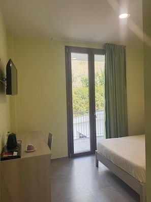Ein Zimmer mit Bett, Schreibtisch, Stuhl, Fenster mit Blick auf Bäume und einem Balkon.