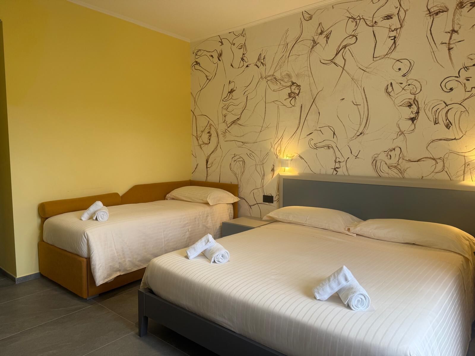A hotel room with two beds, each with a rolled towel, a headboard, and a wall with a line drawing mural.