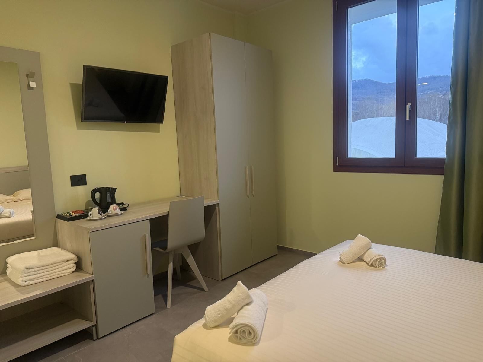 A hotel room with a bed, a desk, a television, a wardrobe, and a window with a view of the outdoors.