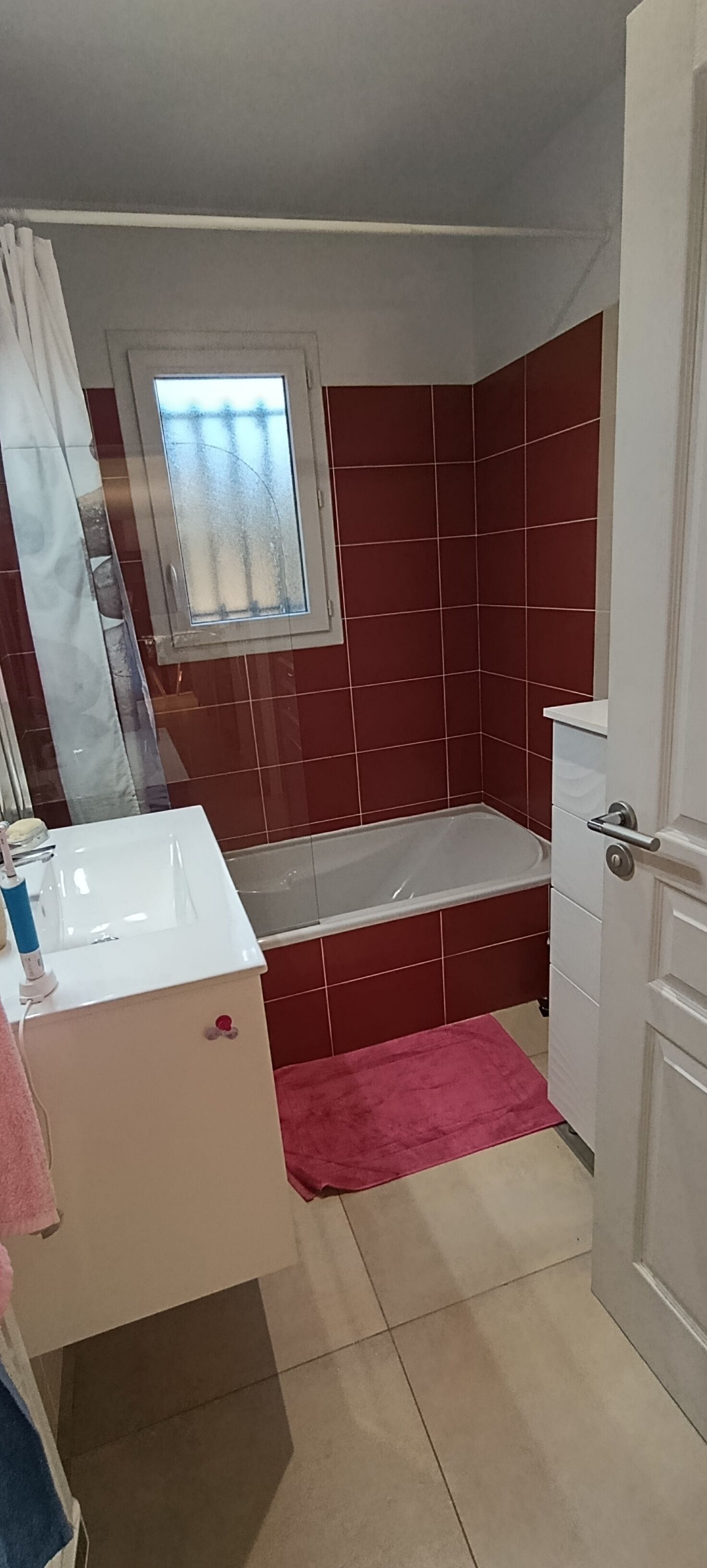 Een kleine, praktische badkamer met een wit wastafel, spiegel, raam en bad.