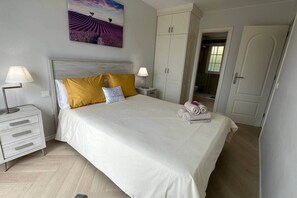 Un letto rifatto con lenzuola bianche e due cuscini, un comodino con una lampada, un armadio e un quadro appeso al muro.
