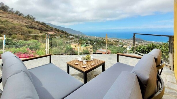 Terraço com duas cadeiras cinza com almofadas e uma mesa de madeira, com vista para uma paisagem costeira com edifícios e vegetação.