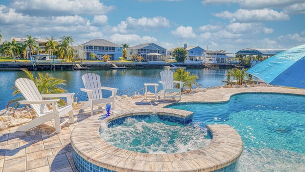 A pool area with a hot tub, lounge chairs, and a view of houses across the water. - NEW! Waterfront Gulf Retreat w/ Heated Pool, Spa, & Mini Golf. (Hernando Beach)