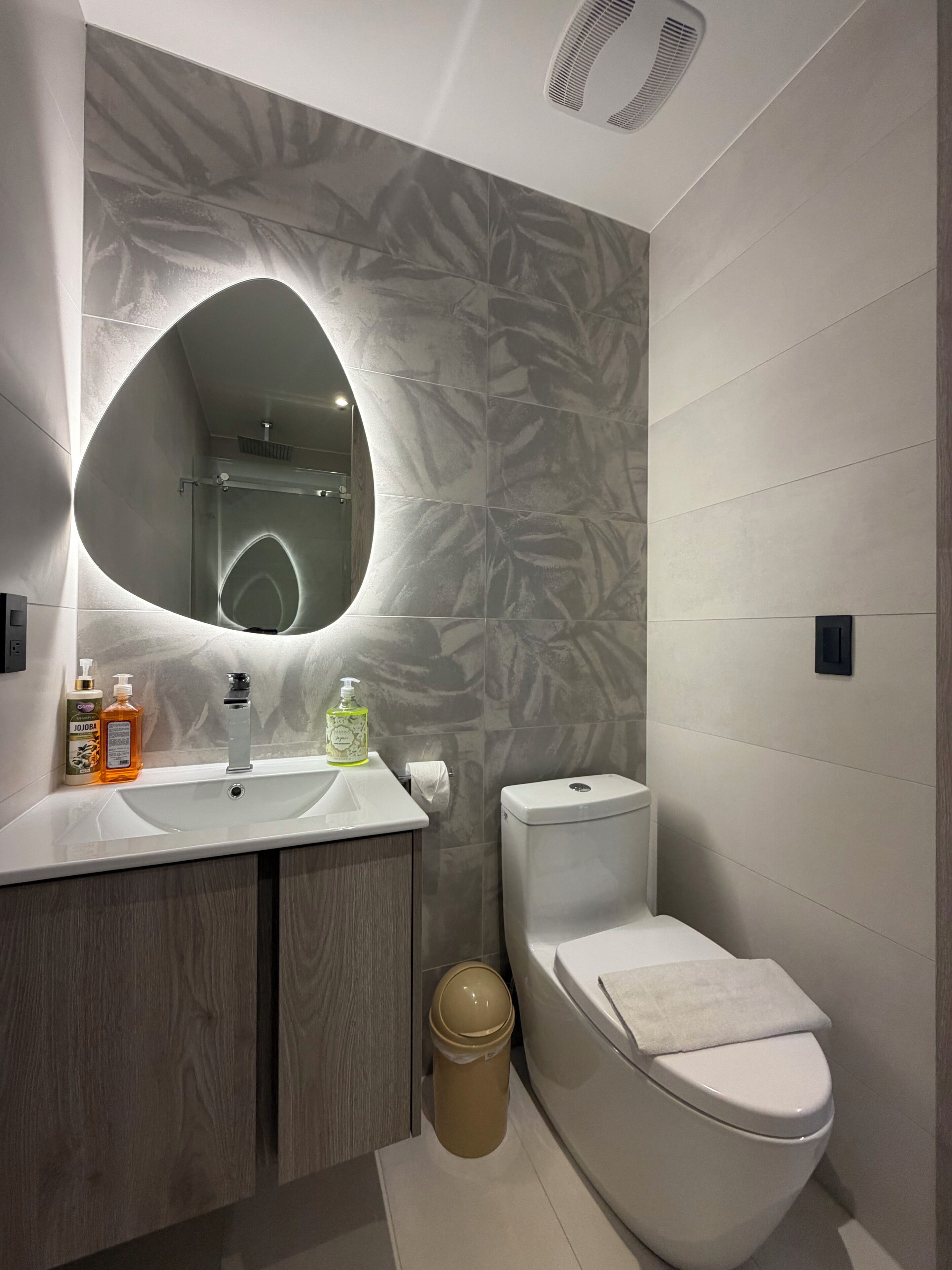 A bathroom with a modern design, featuring a vanity with a mirror, a toilet, and a trash bin.