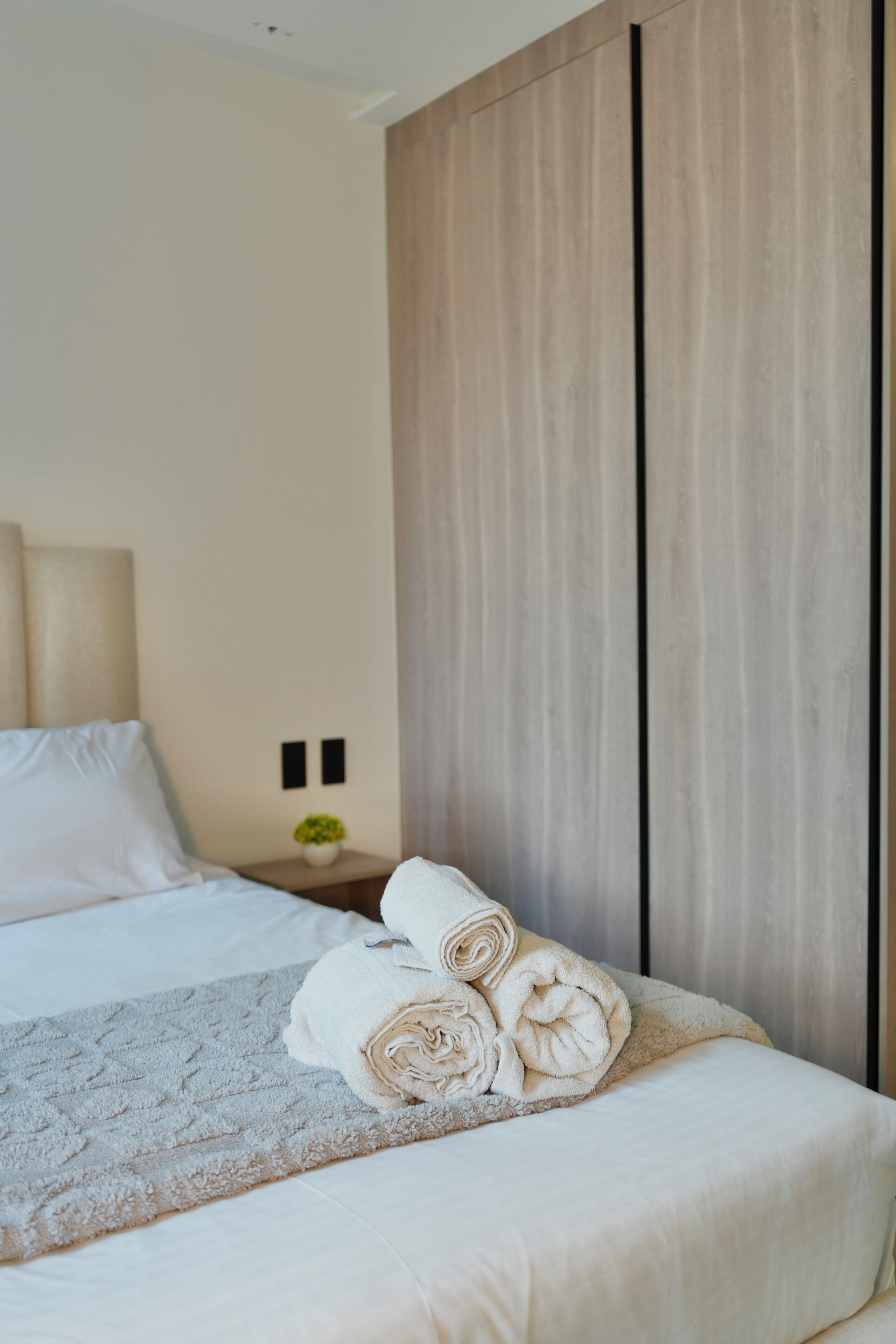 Una cama bien hecha con toallas enrolladas, una mesita de noche con una lámpara y un enchufe en la pared.