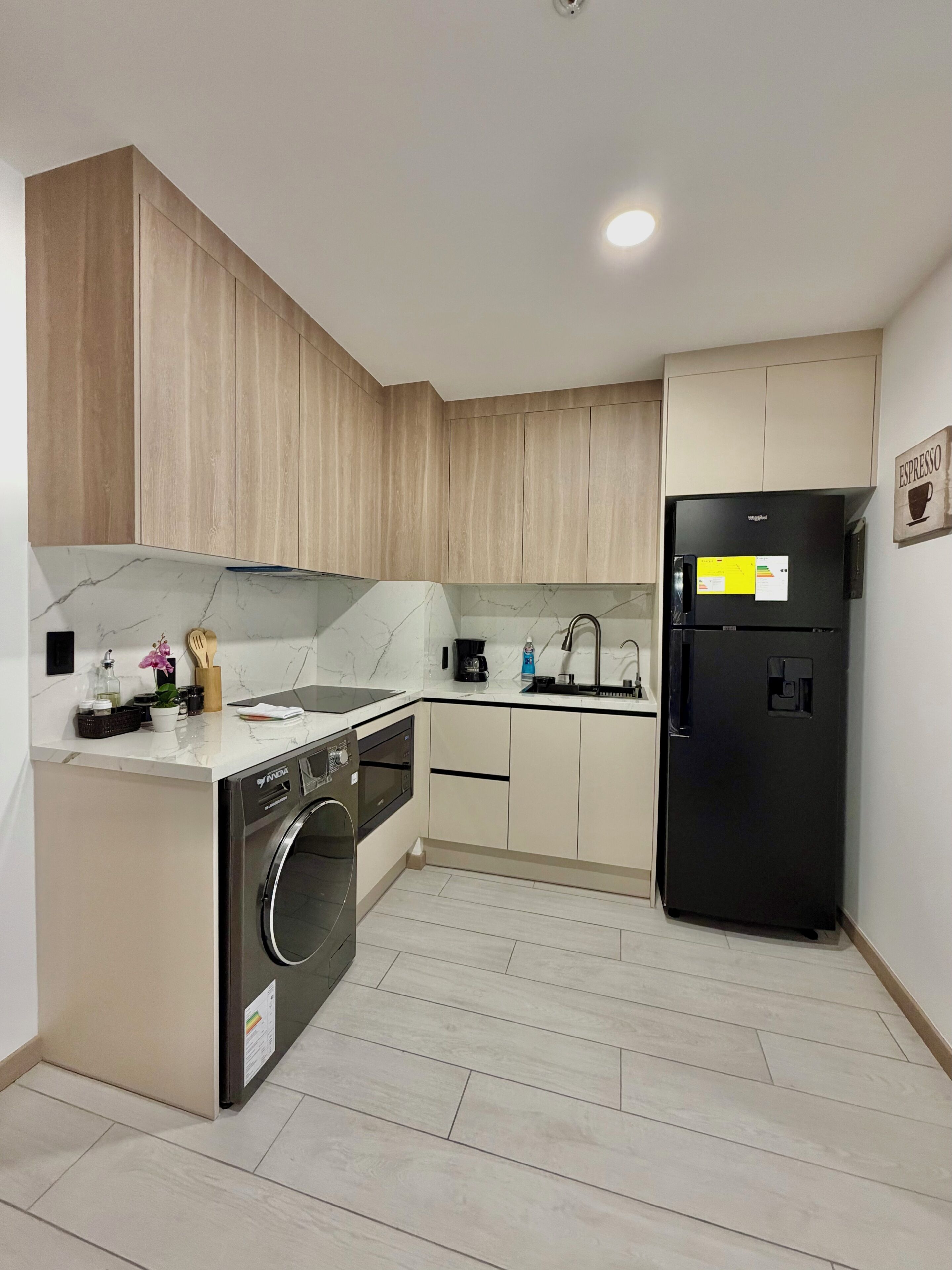 Una cocina compacta con lavadora, un refrigerador negro, armarios de madera y salpicadero de mármol.