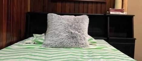 A bed with a green and white striped bedspread, a gray fluffy pillow, and a painting of a forest on the wall behind.