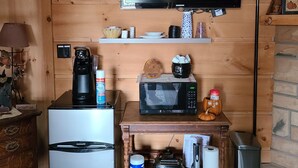 Een ruimtelijk ingerichte leefruimte met een koelkast, magnetron en koffiezetapparaat op een houten tafel. Een televisie is aan de muur boven de tafel bevestigd.