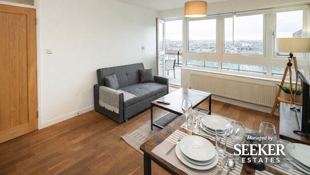 A modern living room with a grey sofa, a dining table set with plates and glasses, and large windows offering a city view. - Shepherd’s Bush • 2 Bed with Terrace • Westfield • (London)