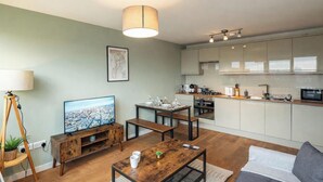 A modern living room with a TV, wooden coffee table, and a sofa. A kitchen with white cabinets and a dining area with a table and chairs is visible. - Shepherd’s Bush • 2 Bed with Terrace • Westfield • (London)