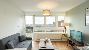 A modern living room with a grey sofa, a coffee table, a TV, and a large window offering a city view. - Shepherd’s Bush • 2 Bed with Terrace • Westfield • (London)