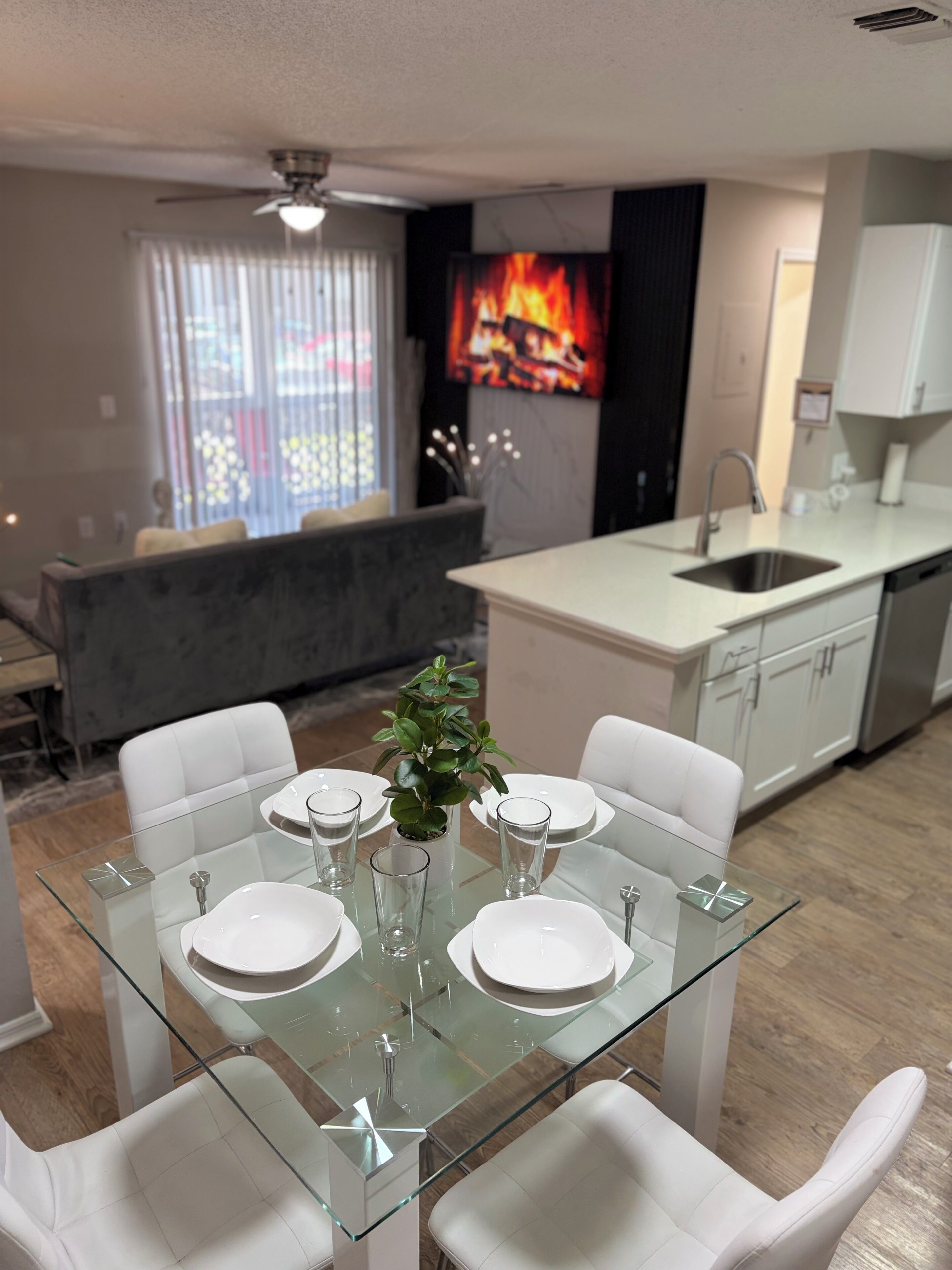 Un comedor moderno con una mesa de vidrio dispuesta para cuatro personas, sillas blancas y una planta en maceta.