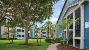A multi-story building with a blue exterior, a covered walkway, and a well-maintained lawn with trees and shrubs. - 2 Bedroom 4 Min to Universal + Free Parking + Fast WiFi (Orlando)