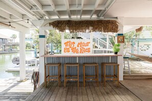 A covered outdoor bar area with a thatched roof, wooden stools, and a sign that reads "THIS MUST BE THE RACE". - Flamingo Way | 2BR + Sleeping Den | Waterfront (Jamaica Beach)