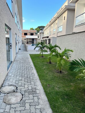 A courtyard with a paved walkway, palm trees, and a pool in the background. - Apartment 13, a 6-minute walk from Taperapuan beach. (Porto Seguro)