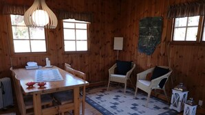 Interior de una cabaña de madera con una mesa de comedor, dos sillas de mimbre, un tapete estampado y una pared de piedra.