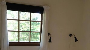 Una habitación con una cama, una mesita con una planta, una ventana con cortinas y dos lámparas fijadas a la pared.