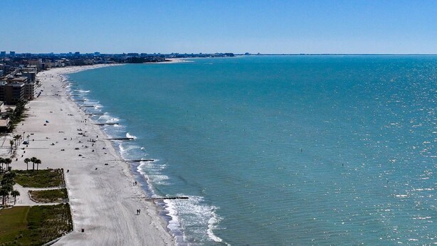 Una spiaggia costiera con un'ampia riva sabbiosa, un mare blu cristallino e uno skyline cittadino in lontananza.