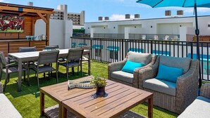 Una terrazza esterna con tavolo, sedie, un divano in vimini con cuscini, un ombrello blu e uno schermo televisivo che mostra una partita di calcio.