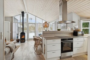 A modern kitchen with a wood-burning stove, white cabinetry, and a large window. - Holiday home with a covered terrace (Ålbæk)