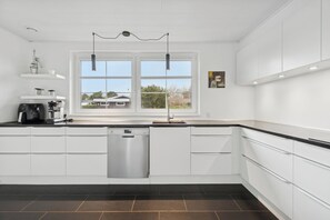 A modern kitchen with white cabinetry, a dishwasher, a coffee machine, and a window with a view of a house. - Relaxing holiday home near beach and harbour (Nexø)