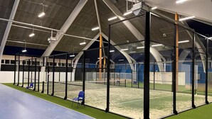En indendørs sportsfacilitet med tennisbane, siddepladser og et hvælvet loft med lysarmaturer.