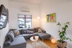A modern living room with a grey sectional sofa, a small white coffee table, a potted plant, and a colorful abstract painting on the wall. - Holiday home in Øer Maritime Ebeltoft (Ebeltoft)