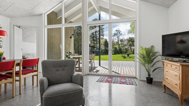 A modern living room with a grey armchair, wooden dining table with red chairs, a television on a wooden cabinet, and a large sliding glass door leading to an outdoor deck. - Cozy little cottage, close to the Limfjord (Storvorde)