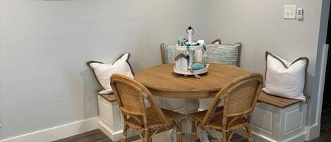 Een eethoek met een ronde tafel, rieten stoelen en een kamerkleed met een patroon. Twee borden aan de muur, waarvan één naar links wijst met het woord 'BEACH' en de andere met de tekst 'zonneschijn, zonsondergang... herhalen'.