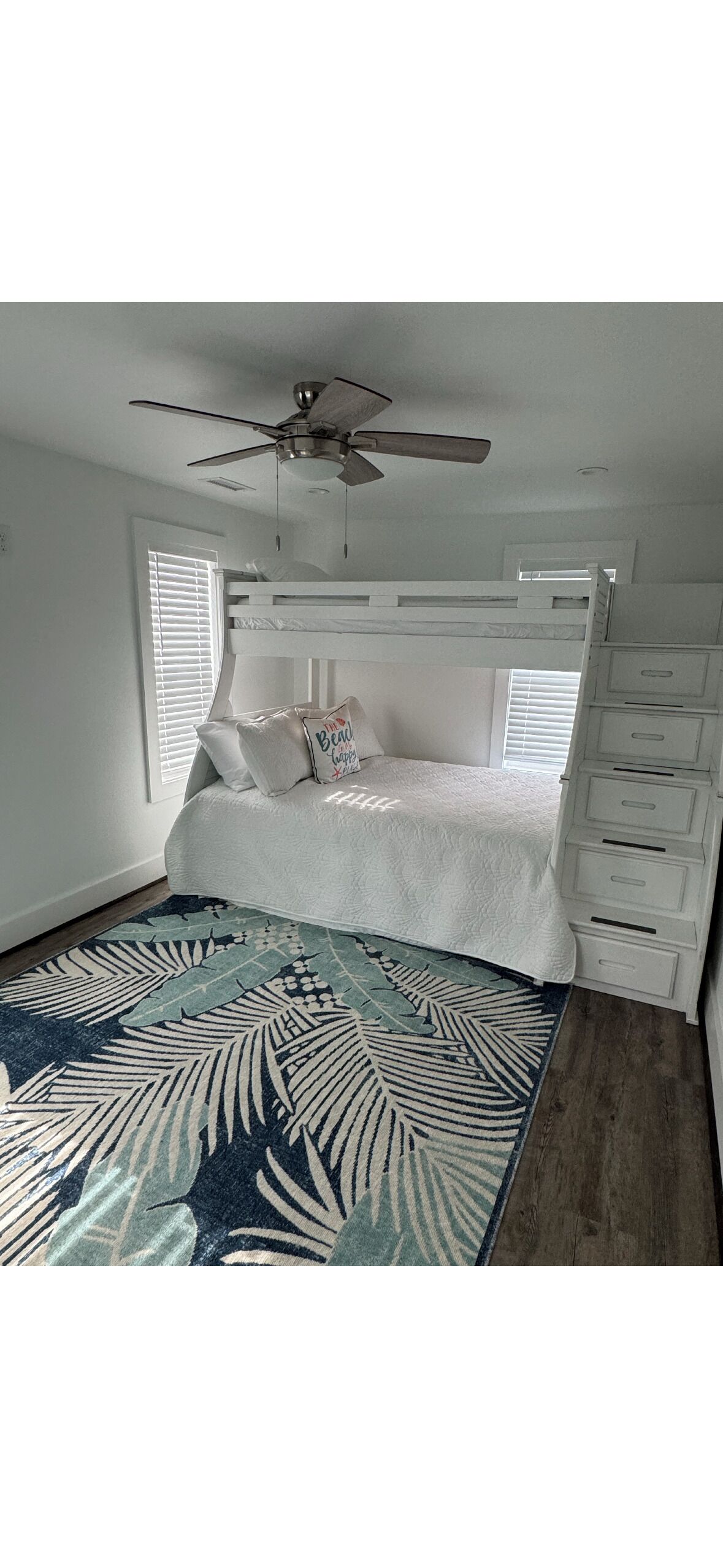 A bedroom with a bunk bed, a ceiling fan, a window with blinds, and a patterned rug.