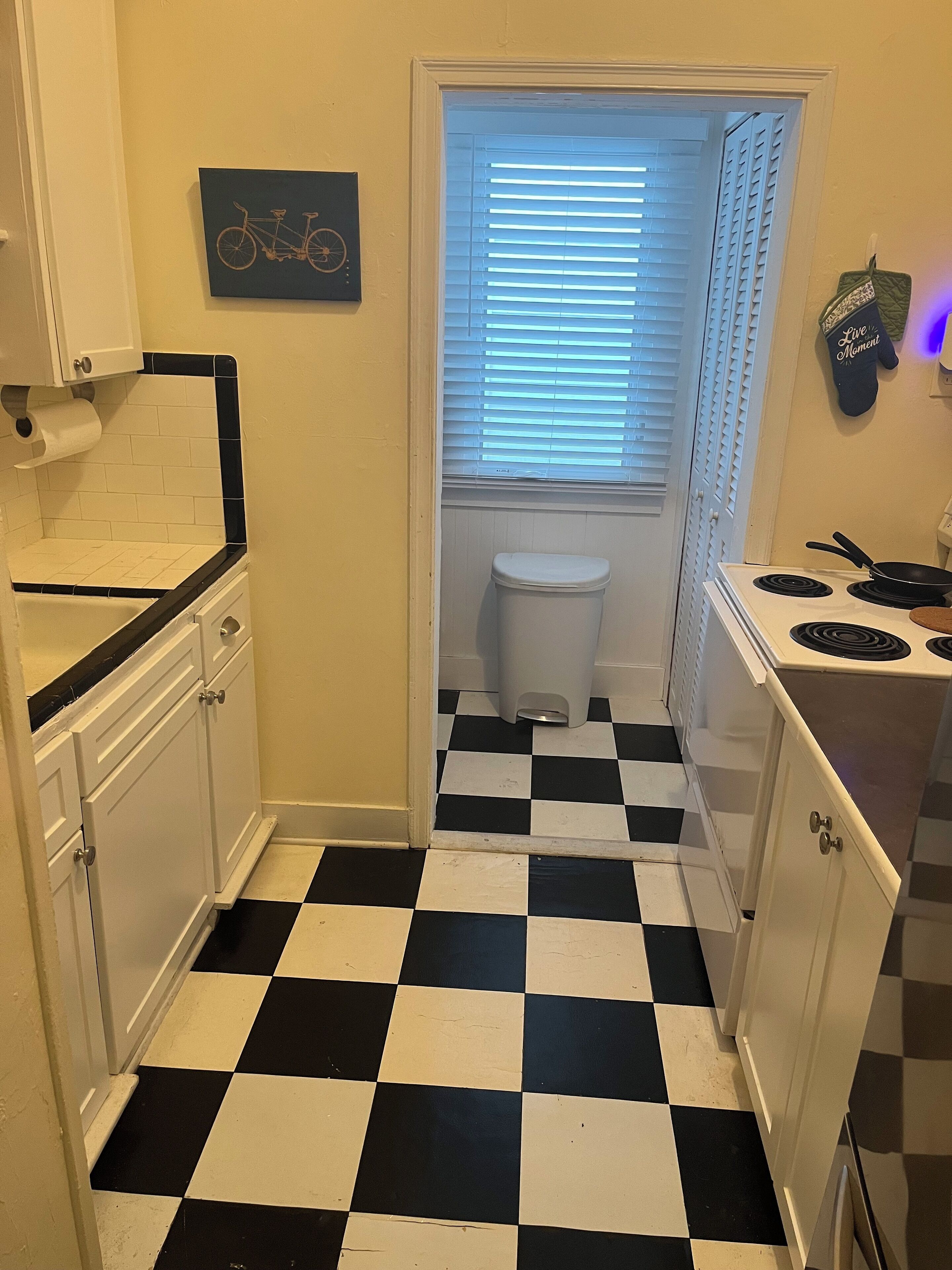 A kitchen with a black and white checkered floor, white cabinets, a stove, and a window with blinds.