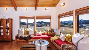 A cozy living room with a red sofa, multiple cushions, a glass coffee table, and a wooden cabinet with dishes. Large windows offer a view of the outdoors. - Cozy 1BD & Loft for 4 Near Community Pool & Trails (Sea Ranch)