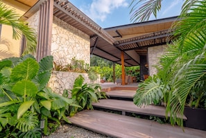 Un espace extérieur abrité, doté d’un mur en pierre, d’escaliers en bois et de plantes tropicales.