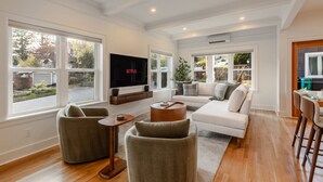 A spacious living room with a large flat-screen TV, comfortable seating, and large windows offering a view of the outdoors. - Phinney Ridge Luxe Retreat | Gym, BBQ & Game Room (Seattle)