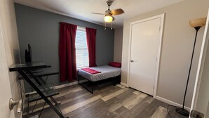 Une petite pièce comprenant un lit, un bureau, un ventilateur de plafond et une porte.