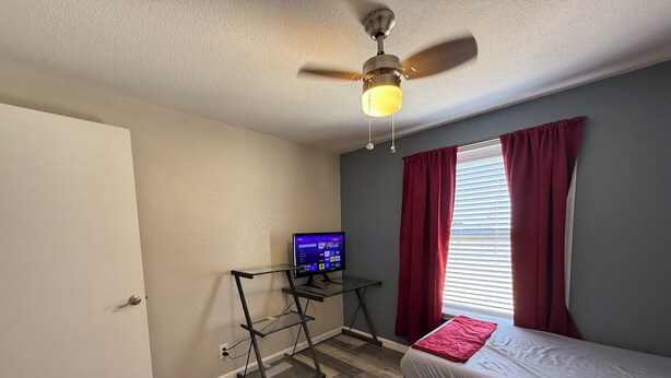 Une chambre à coucher avec un lit, un bureau avec un ordinateur, un ventilateur de plafond et une fenêtre avec des rideaux.