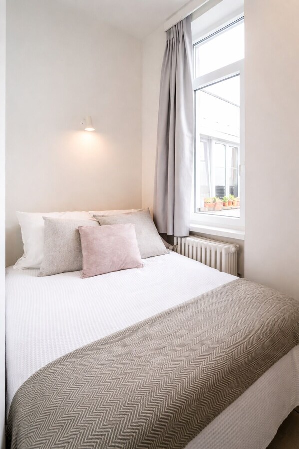A neatly made bed with a patterned blanket and pillows, a radiator, and a window with curtains. - Antwerp Short Stay Apartments (Antwerp)