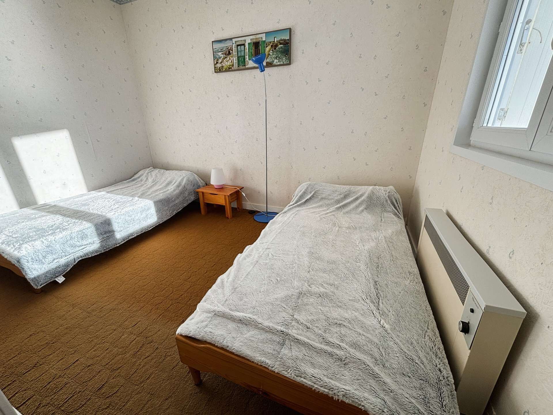 A room with two single beds, a small table with a lamp, a picture on the wall, and a window.