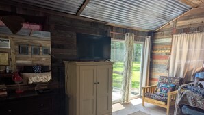 Ein gemütliches Zimmer mit Holzbalkendecke, einem großen Fernseher auf einer Kommode, einem Bett mit gemusterten Betttextilien, einem Sessel mit bunten Kissen und Blick auf eine grüne Landschaft durch eine Schiebetür.