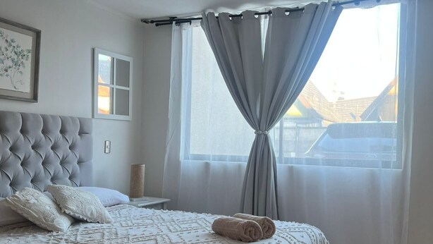A bedroom with a tufted headboard, a bed with a patterned bedspread, a bedside table with a lamp, a window with curtains, and a ceiling light fixture. - Brisa del Sol Apartment - Comfortable and well located (Talcahuano)
