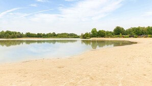 Una playa arenosa con un lago tranquilo, rodeada de árboles y algunos edificios al fondo.