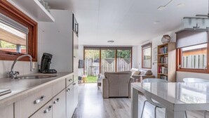 Una cocina moderna con estufa integrada, cafetera y lavaplatos. Hay un comedor con mesa y sillas, y una estantería llena de libros.