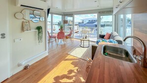A modern kitchen with a wooden countertop and a sink, a dining area with a table and chairs, and large windows offering a view of boats on the water.