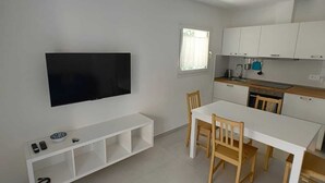 Een moderne keuken met witte kastjes, een eettafel met stoelen, een aan de muur bevestigde televisie en een klein raam.