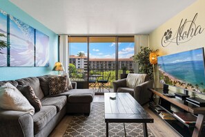 Ein Wohnzimmer mit Sofa, Sessel, Couchtisch und Fernseher. An den Wänden befinden sich Gemälde, eine Pflanze und ein Blick auf den Ozean durch große Fenster.